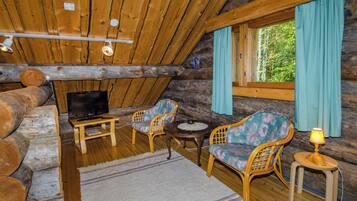 Interior de cabana de madeira com televisão, duas cadeiras de vime, uma mesa pequena e uma janela com cortinas.
