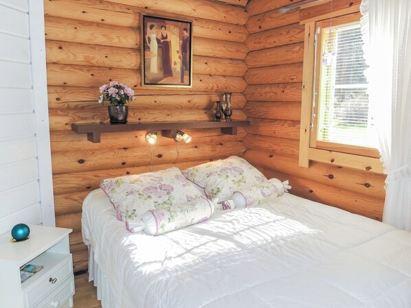 Un dormitorio de cabaña de madera con una cama, una mesita de noche, un aparador, una estantería con objetos decorativos y una ventana con cortinas.