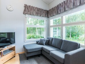 Een moderne woonkamer met een grijze modulaire bank, een flatscreen televisie op een houten dressoir en een groot raam met uitzicht op groen.