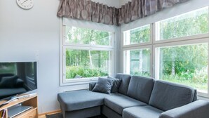 A modern living room with a grey sectional sofa, a flat-screen TV on a wooden stand, and large windows offering a view of greenery. - Prokatti by Interhome (Sotkamo)