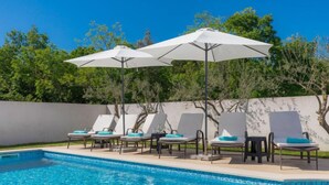 Piscina con sillas de descanso y sombrillas, rodeada por un muro bajo y árboles.