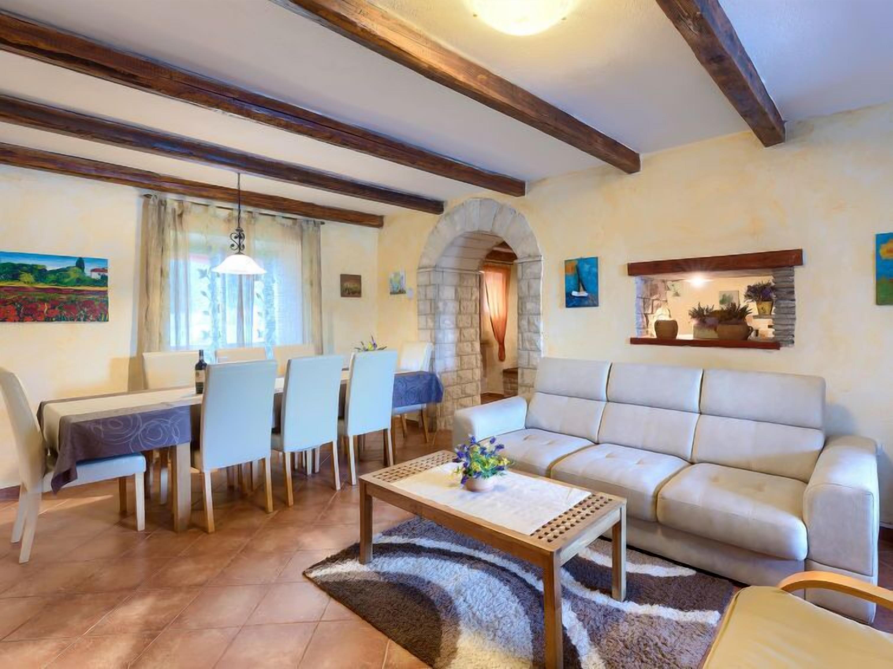 A living room with a sofa, dining table, chairs, and a coffee table. There is a stone archway and a shelf with plants.