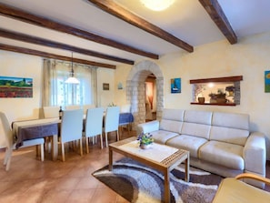 Un salon comprenant un canapé, une table à manger, des chaises et une table basse. On y trouve une arche en pierre et une étagère ornée de plantes.