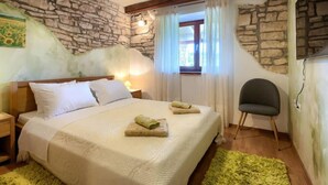 Un dormitorio con pared de piedra, cama con ropa blanca, cabecera de madera, una silla verde y una ventana con cortinas.