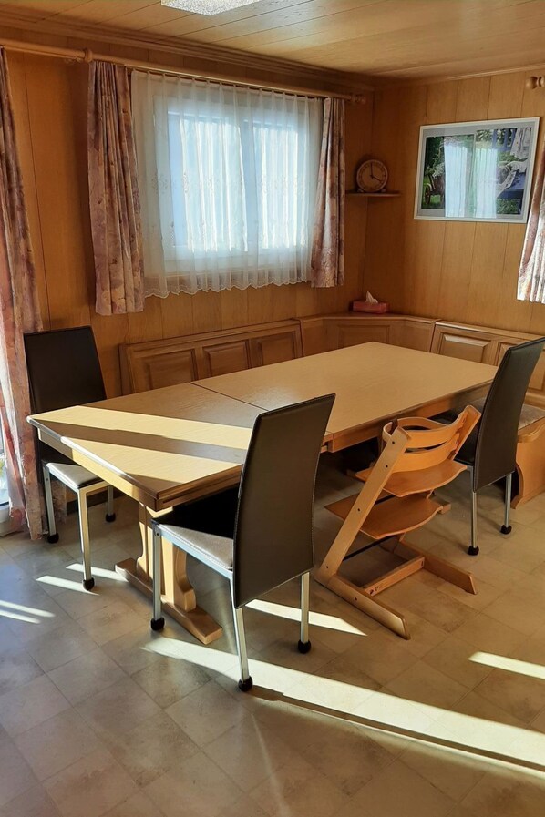 Ein Essbereich mit einem großen rechteckigen Tisch, sechs Stühlen und einem Fenster mit durchsichtigen Vorhängen.