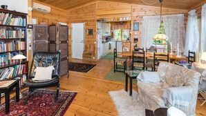 A cozy cabin interior with a wooden ceiling, a living room with a sofa and chairs, a dining area, a kitchen, and a bookshelf filled with books. - Metsä-iivari by Interhome (Lohja)