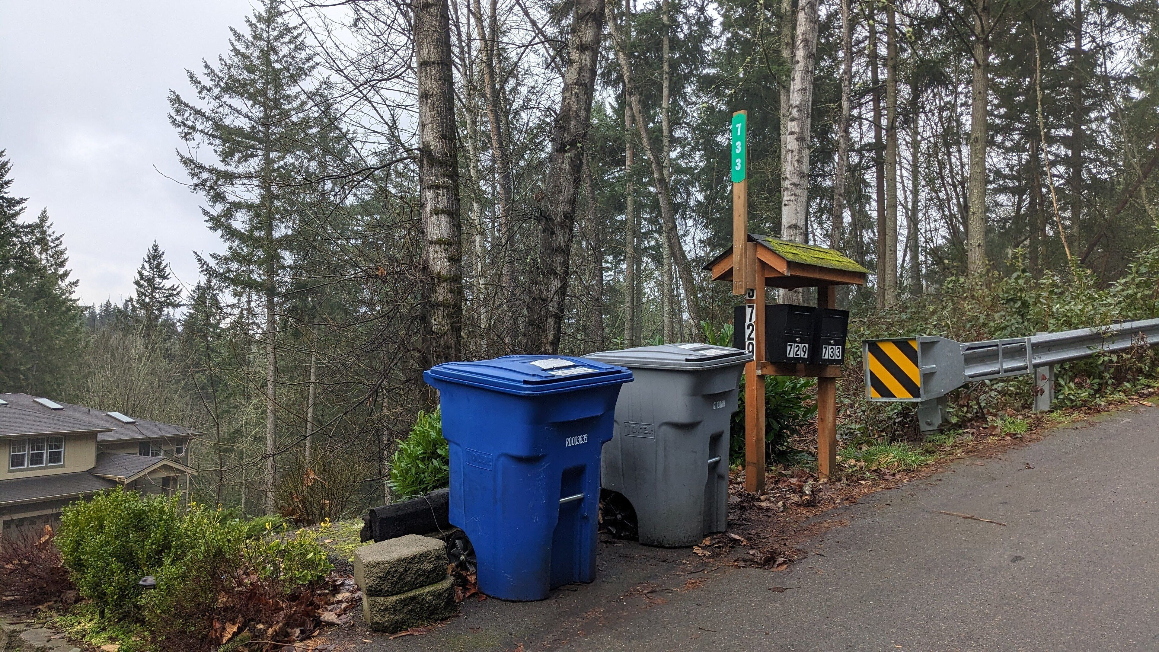 A roadside with a wooden signpost, two trash bins, and a guardrail.