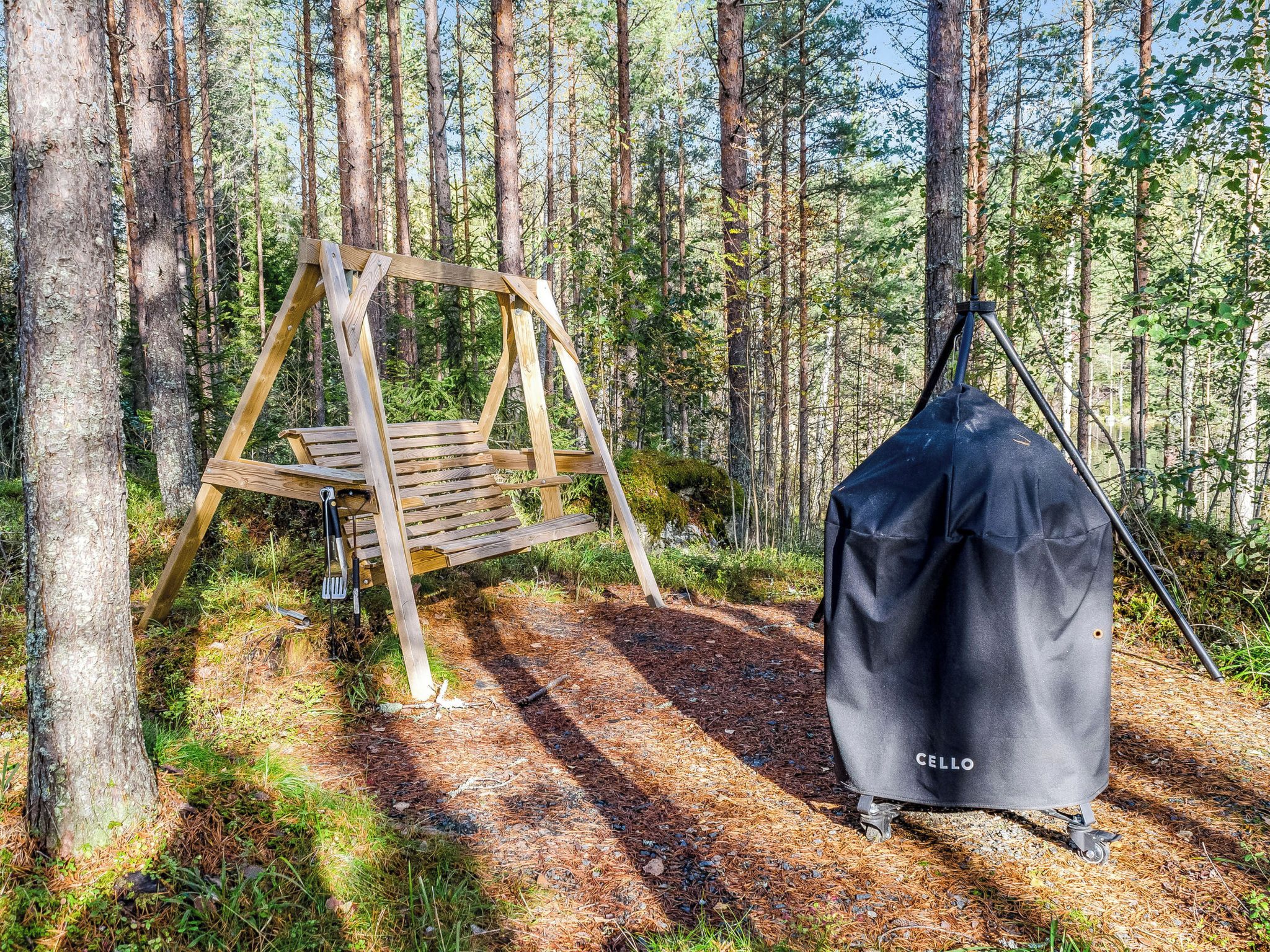 Ein hölzernes Schaukelgestell in einer bewaldeten Gegend mit einem schwarzen Grillzelt mit der Aufschrift 'CELLO' in der Nähe.
