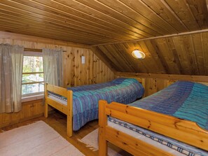 Una cabana de fusta amb dos llits de fusta, una finestra amb cortines, i un sostre inclinat amb una làmpada.