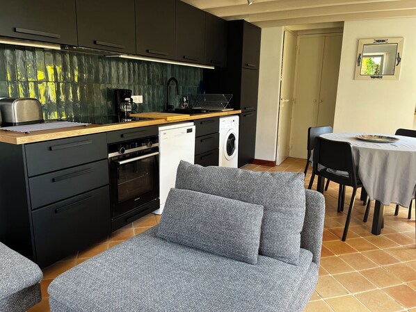 A compact kitchen with modern appliances, a dining area with a table and chairs, and a grey sofa.