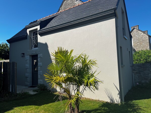 A house with a stone facade, a dark roof, and a small garden with palm trees.