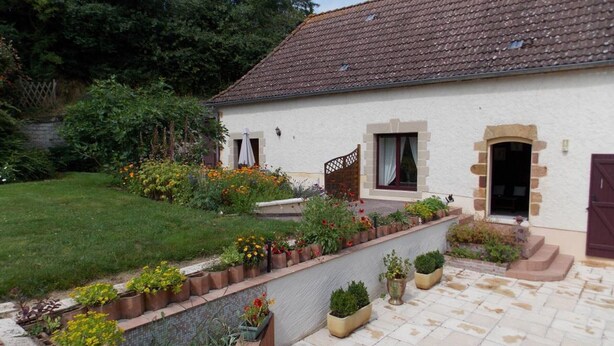 A white house with a tiled roof, a garden with various plants and flowers, and a paved patio area.
