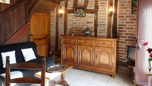 A rustic room with exposed brick walls, wooden beams, a wooden cabinet with a dish on top, a wooden chair, and a black sofa with white cushions.