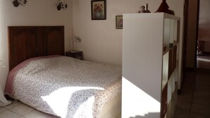A bedroom with a wooden headboard, a bed with a floral bedspread, a nightstand with a lamp, and a white wardrobe.