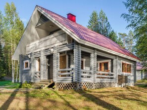 Hirsimökki, jossa on jyrkkästi kallistuva katto, kuisti kaiteineen ja savupiippu.