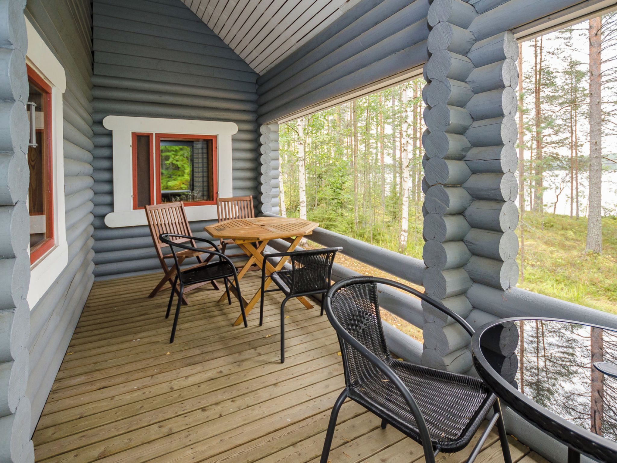 A log cabin with a wooden deck, a table with chairs, and a window overlooking a forest.