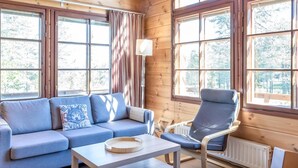 Interior acogedor de una cabaña con un sofá azul, una silla y una mesa. Amplias ventanas ofrecen luz natural y vistas al exterior.