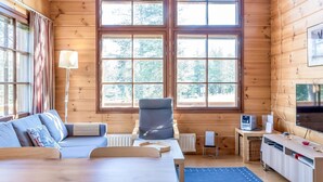 Un interior de cabaña acogedor con un ventanal grande, un sofá, una silla, un escritorio y un televisor.