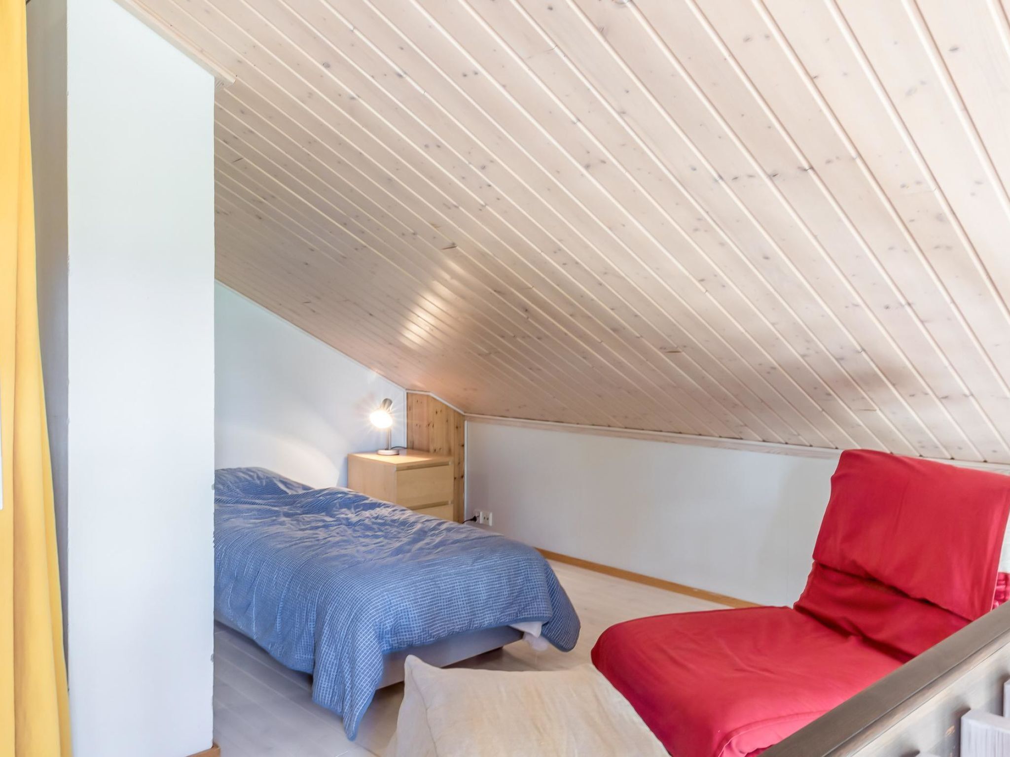 A bedroom with a sloped ceiling, a bed with a blue and white checkered duvet, a red chair, and a bedside table with a lamp.