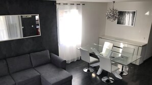 A modern living room with a grey sofa, a glass dining table with white chairs, a small white coffee table, a black-framed mirror, and a chandelier. - Chalet in Provence with Pool, Parking, Wifi, Near Avignon, Luberon ... (Sorgues)