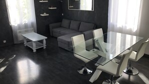 A modern living room with a glass dining table, white chairs, a grey sofa, and a small white side table. - Chalet in Provence with Pool, Parking, Wifi, Near Avignon, Luberon ... (Sorgues)