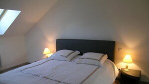 Un lit bien fait, avec une tête de lit sombre, dans une pièce aux plafonds inclinés et dotée d’un puits de lumière. Deux lampes de chevet sont placées de part et d’autre du lit.