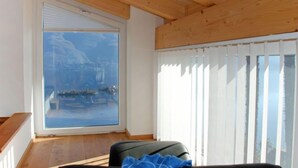 Ett rum med ett stort fönster som ger utsikt över en sjö eller ett hav och berg, ett träpanelat tak och en svart soffa med en blå handduk.
