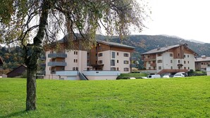 Un immeuble résidentiel doté de nombreux balcons, un champ verdoyant au premier plan et un paysage montagneux en arrière-plan.