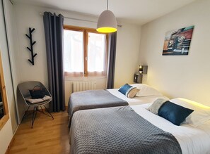 A hotel room with two beds, a chair, a radiator, a window with curtains, a wall clock, and a painting.