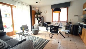 Un salon moderne comprenant un canapé, une table basse et un coin repas avec des chaises et une table. On y trouve également une bibliothèque, une plante en pot et un coin cuisine avec des placards et un évier.