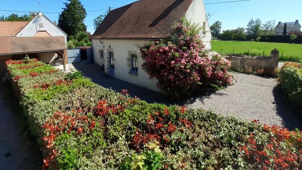 A white house with a brown roof, surrounded by a gravel driveway and flowering plants.