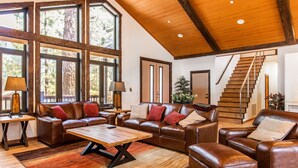 A spacious living room with leather sofas, a wooden coffee table, and large windows offering a view of trees. A wooden staircase is visible in the background.