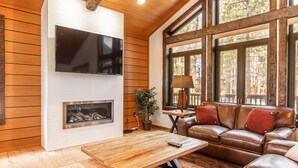A modern living room with a fireplace, leather sofa, and large windows offering a view of trees.