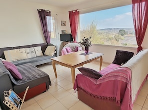 Sala de estar com sofá, mesa e televisão. Janela com vista para o campo.