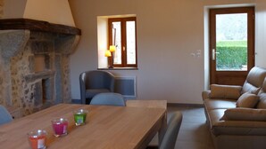 Un salon chaleureux avec une cheminée en pierre, une table à manger en bois décorée de bougies, un canapé confortable et une fenêtre donnant sur un paysage verdoyant.