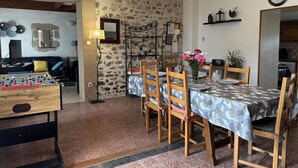 Ein gemütliches Wohnzimmer mit einem Kicker, einer Couch und einem Couchtisch. Es gibt einen Essbereich mit einem Tisch für zwei Personen, Stühlen und einer Blumenvase. Eine Uhr hängt an der Wand, und hinter dem Essbereich ist eine Steinwand zu sehen.