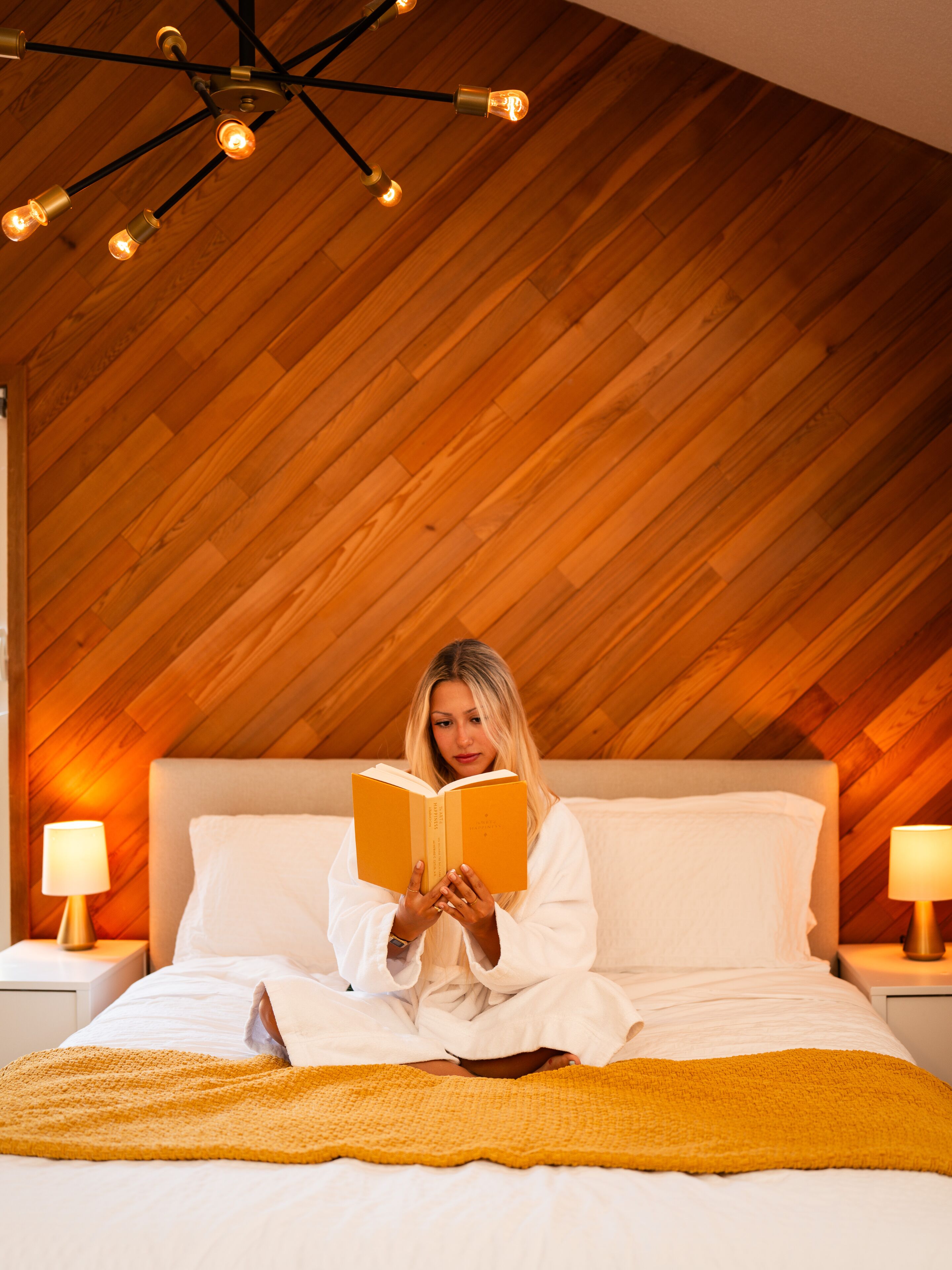 A person sitting on a bed in a room with wooden slats on the ceiling and two bedside lamps.