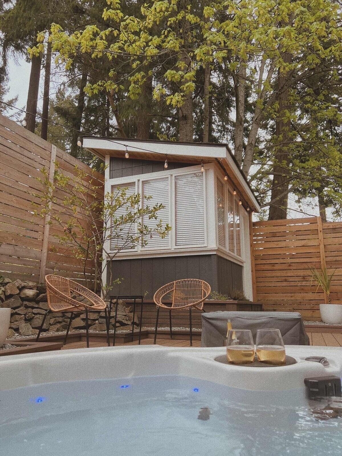 Eine kleine Holzhütte mit Whirlpool, zwei Korbstühlen und einem Tisch mit Gläsern und Flaschen.