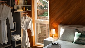 A bedroom with a wooden ceiling, a bed with white bedding, a bedside table with a lamp, a window with a view of greenery, and a bookshelf with books and a robe rack. - ★Ocean View Loft w/ Dedicated Hot Tub in Departure Bay. Treat Yourself Book Now★ (Nanaimo)