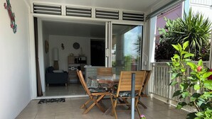 A patio with a dining table and chairs, a sliding glass door leading to a living area, and a garden with flowers. - ■ BEAUTIFUL T2 CLIMATISE IN VILLA SEA VIEW - RAVINALA MARTINIQUE ■ (LES TROIS ILETS)