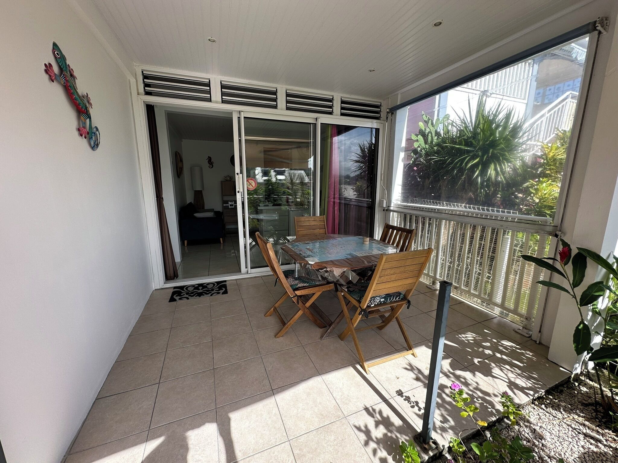 A sunlit patio with a table and chairs, a sliding glass door leading to a balcony, and a view of greenery outside.