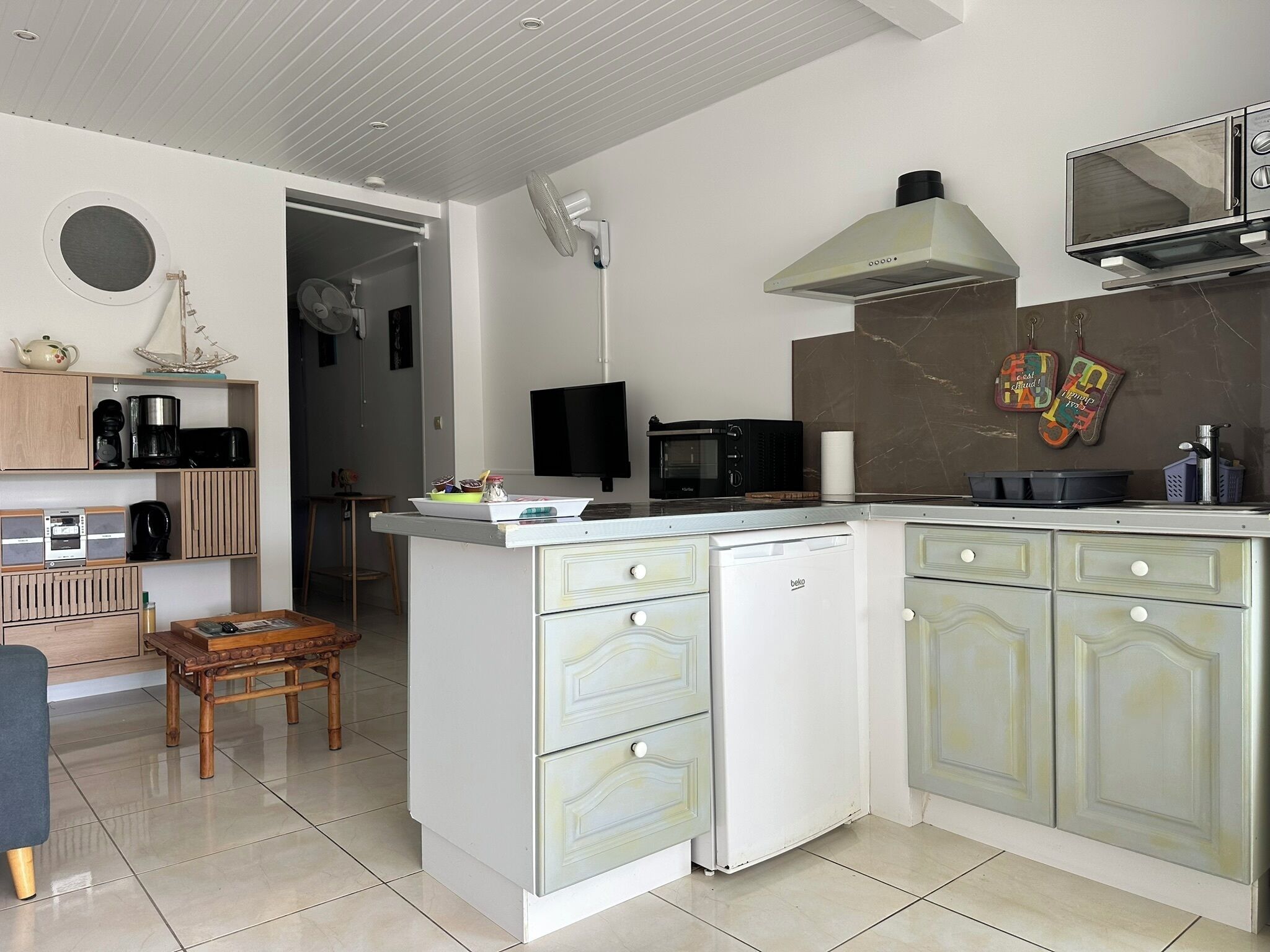 A compact kitchen with a built-in microwave, a small refrigerator, and a sink. There is a wooden dining table and a small stool in the dining area.