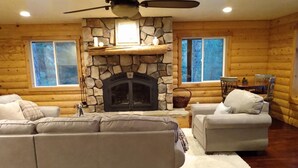 Ein gemütliches Wohnzimmer mit Kamin aus Stein, zwei Sofas, einem weißen Teppich und einem Essbereich mit Tisch und Stühlen.