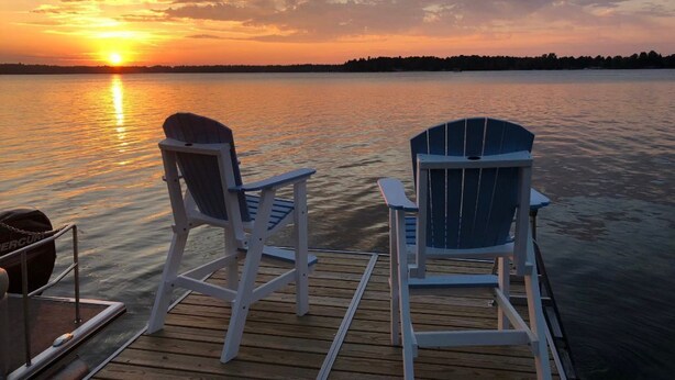 Twee Adirondack stoelen op een houten steiger, met een zonsondergang boven een rustig meer.