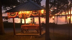 Een kampvuur met een houten tafel en banken onder een paviljoen met Amerikaanse vlaggen, met uitzicht op een waterpartij.