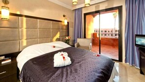 Hotellihuone, jossa on suuri sänky, sängynpääty, televisio, yöpöytä ja ikkunasta avautuva näkymä rakennukseen.