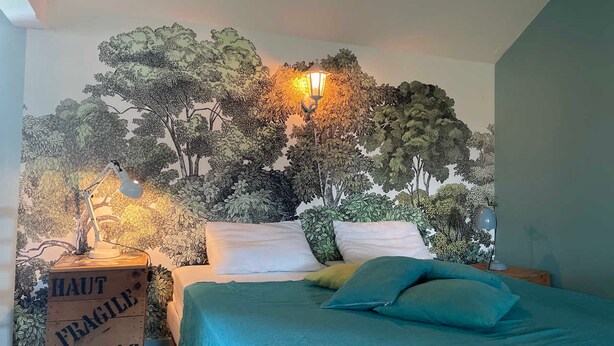 A bedroom with a mural of trees on the wall, a bed with teal bedding, and a nightstand with a lamp.