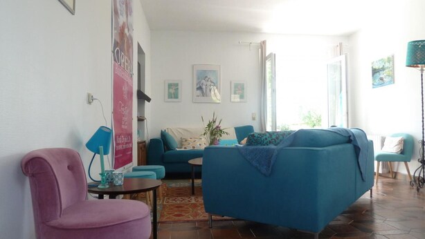 A living room with a blue sofa, a pink chair, and a small table with a lamp. There are framed pictures on the wall and a window with curtains. - Country house with enclosed garden - Pets welcome (Barrais-Bussolles)