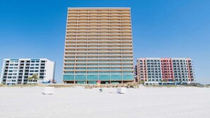 A tall hotel with multiple floors and balconies, situated on a sandy beach with other buildings nearby.
