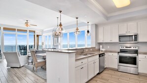 Une cuisine moderne avec des meubles blancs, des appareils en acier inoxydable et un grand îlot central. Elle offre une vue sur l’océan grâce à de grandes fenêtres.